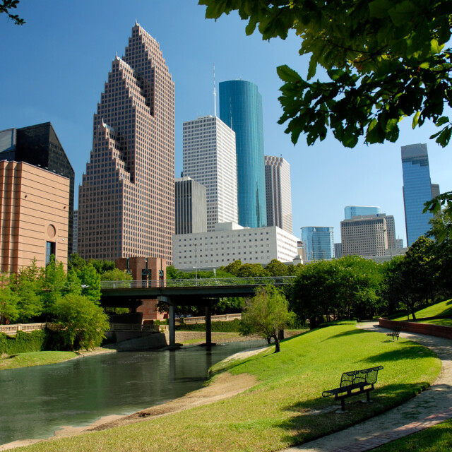 Modern skyscrapers of Houston, Texas are seen in this horizontal color image across a waterway flowing through a city park. A perfect, clear blue sky is a backdrop to this afternoon cityscape. Tops of the buildings are somewhat outlined by tree branches. A park bench and walking trail are included in the foreground.